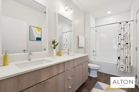 Modern apartment bathroom with double vanity, mirrors, white tile bathtub, and wood cabinets.
