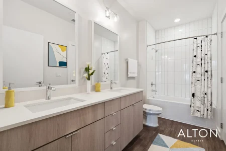 Modern apartment bathroom with double vanity, mirrors, white tile bathtub, and wood cabinets.