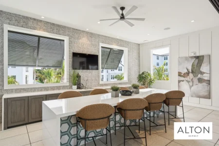 Apartment clubhouse with large kitchen island, bar stools, wall-mounted TV, and windows.